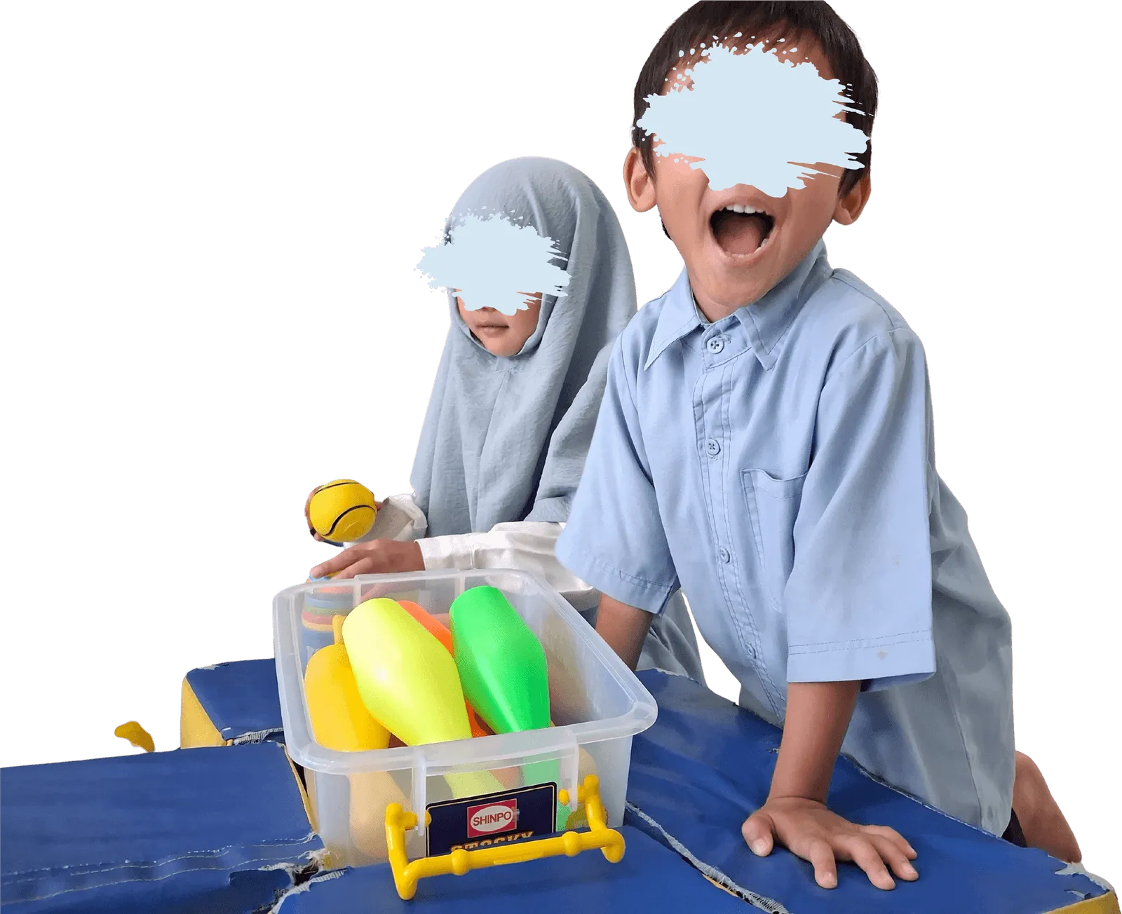 Dua anak Prasekolah berseragam biru muda di atas matras, satu tertawa di samping kotak berisi mainan